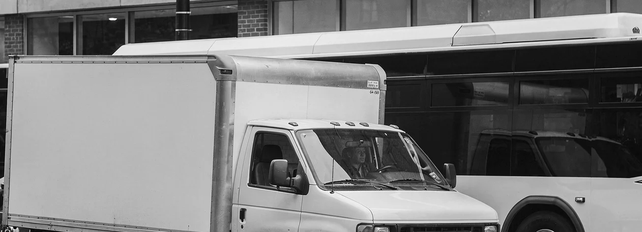 A white delivery truck double-parked at a retail storefront curb, a city bus stopped directly behind it, blocked from passing.