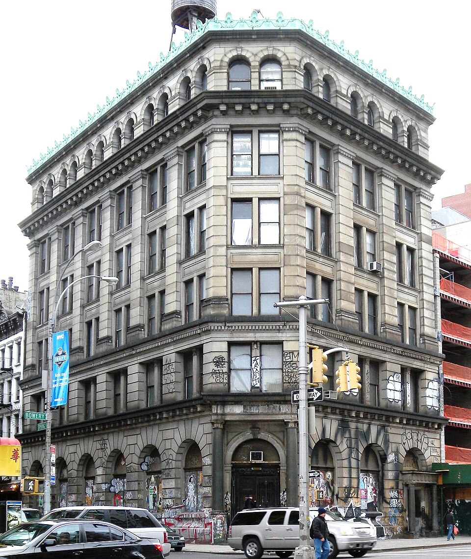 Corner view of 190 Bowery's stone facade, graffiti-covered lower floors, traffic, and the Bowery street sign.