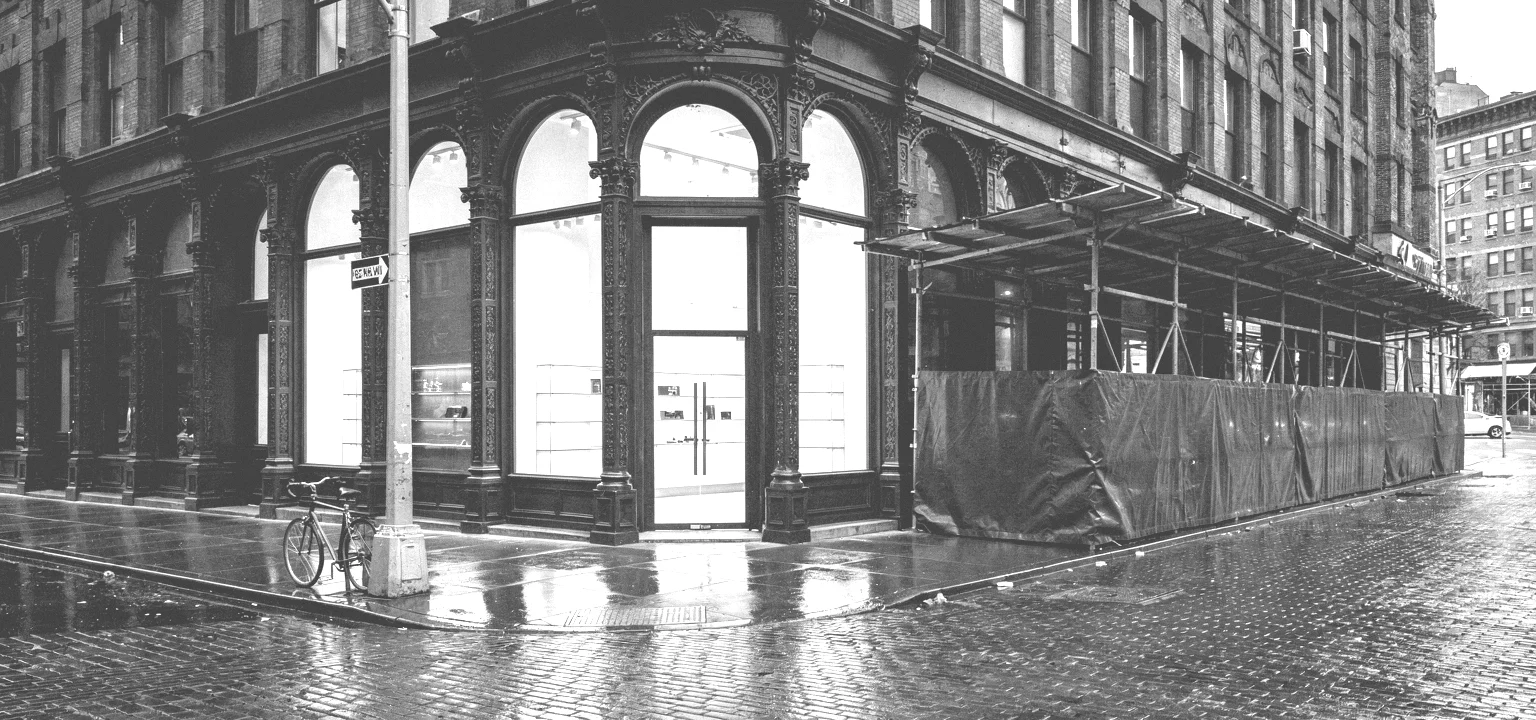 A rain-soaked SoHo cast-iron corner with a glowing retail storefront beside a scaffolding shed.