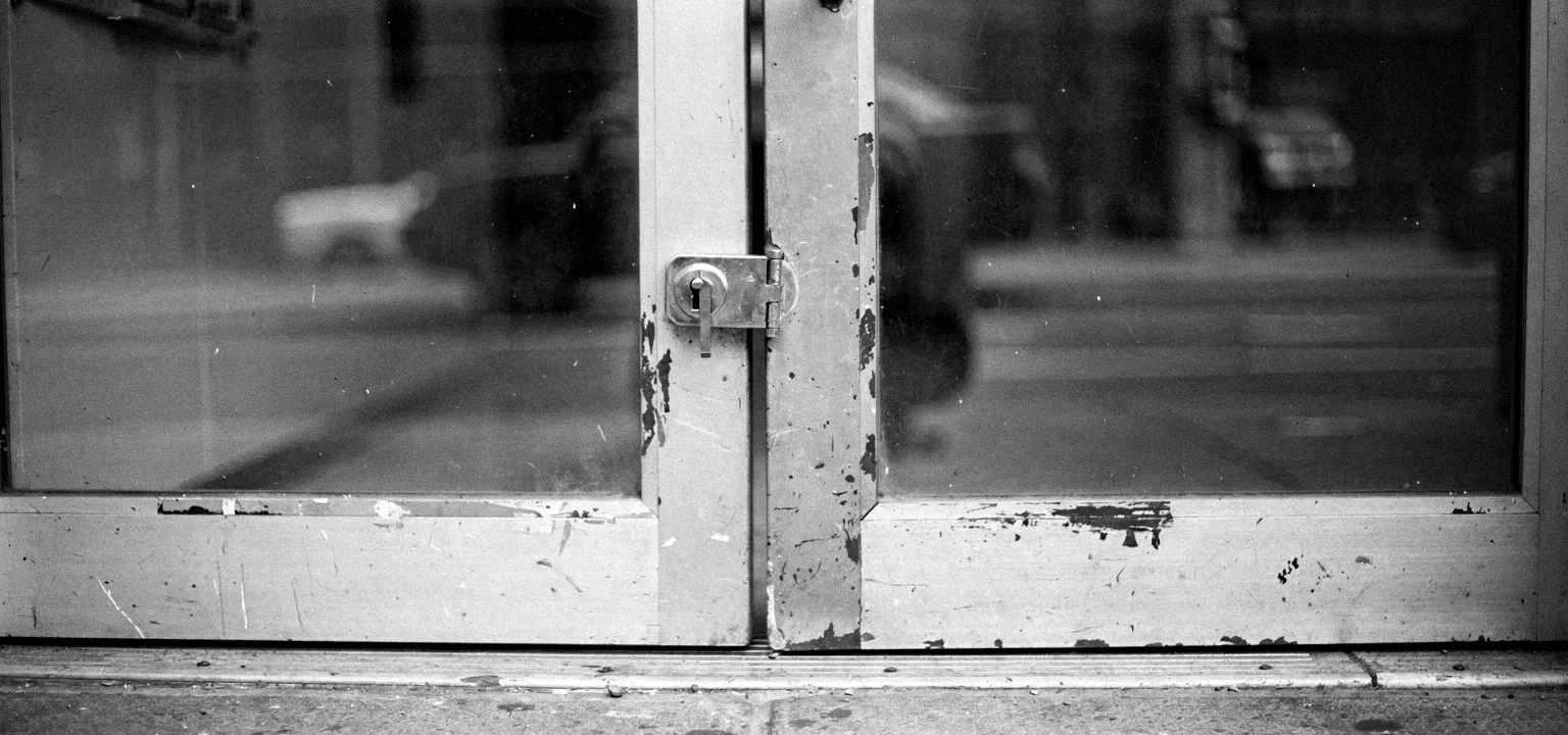 A closed storefront glass door with a heavy deadbolt and keyhole, the question of who holds the keys.