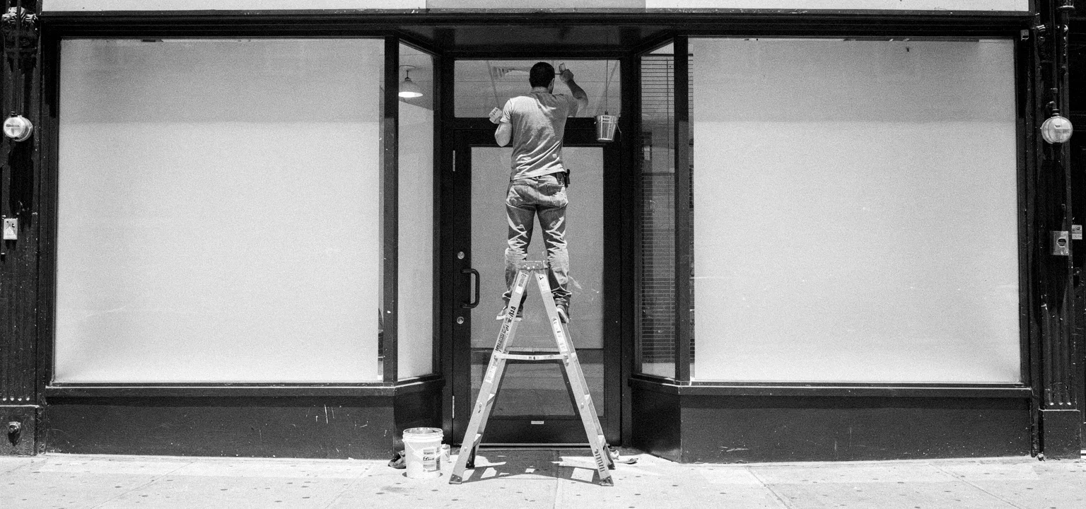 A worker on a ladder repainting a blank storefront in daylight.
